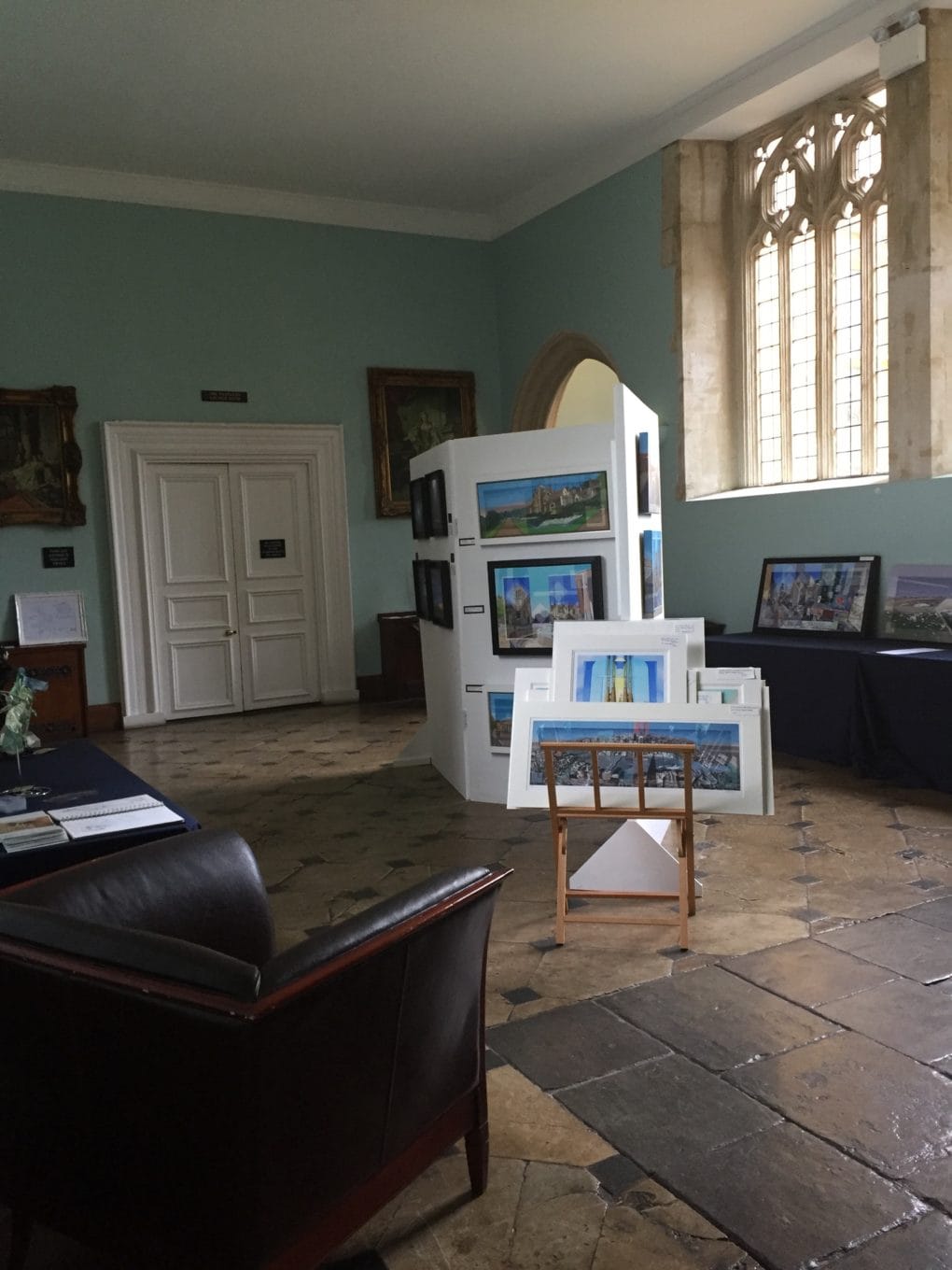 A view of the Great Hall Gallery facing onto the West Lawn. In front is a chair, a stand with prints and a temporary wall displaying artworks by Suzy Davis and Len Matthews. 