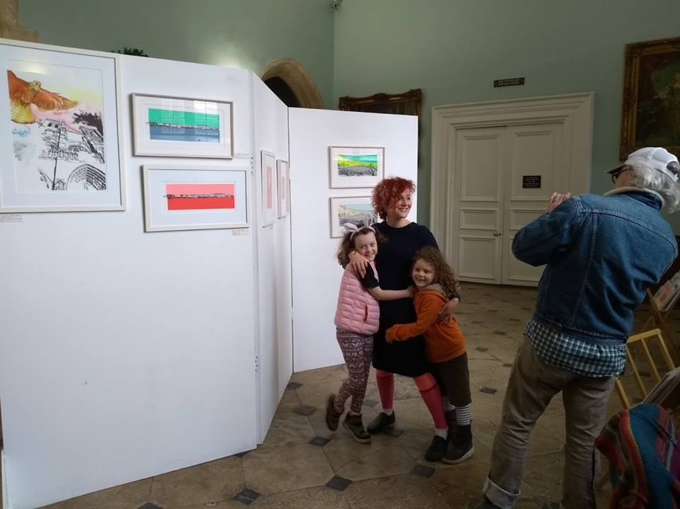 Amy Hutching exhibition launch. Amy is standing in front of her exhibition and being hugged by her two daughters. Her husband is taking a photograph. 