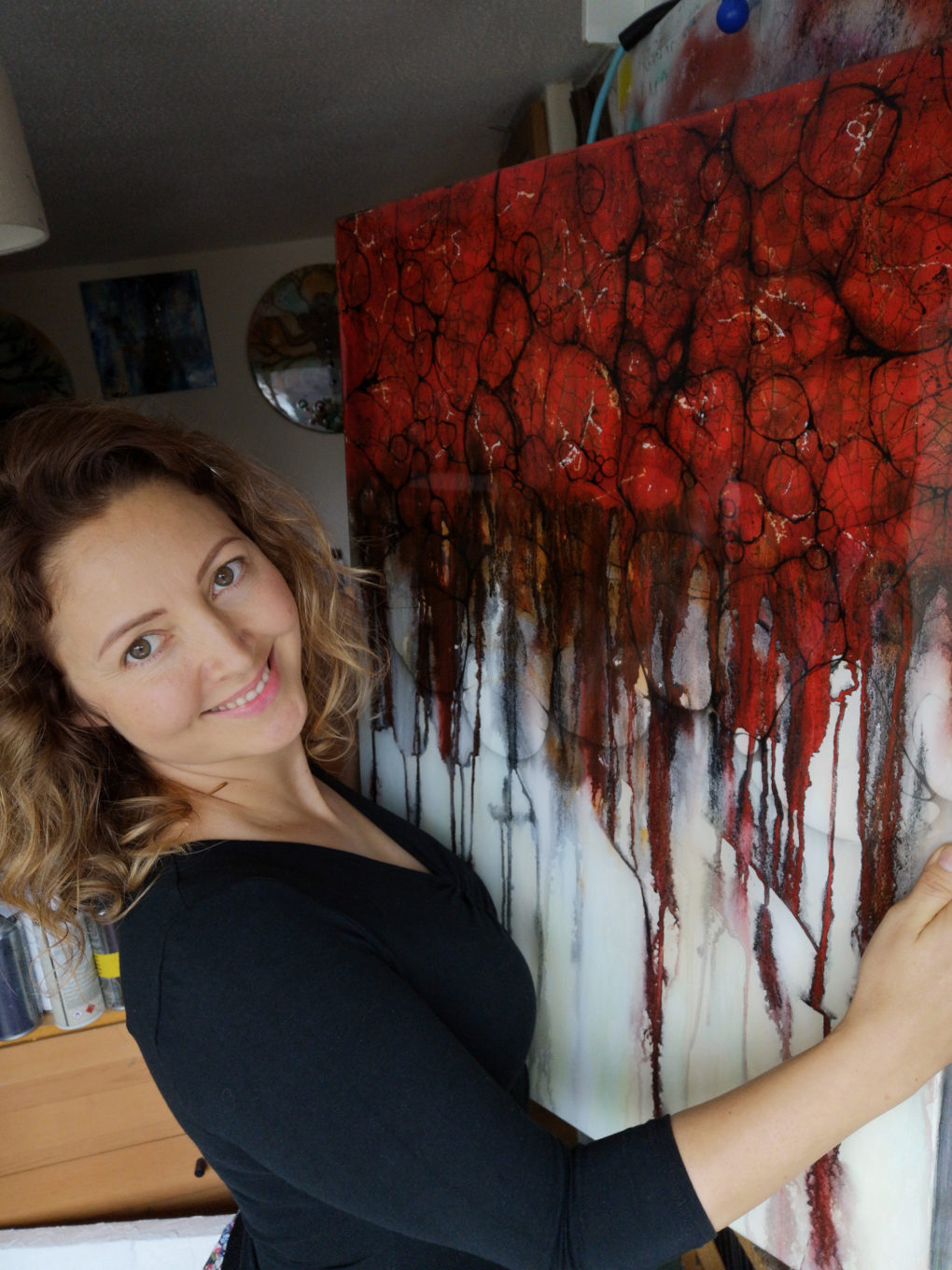 Jeanette is a white woman. She has mid length curls and is turned to face the camera and smiling. She is wearing a black top and is holding a painting which has vivid red circles and lines. 