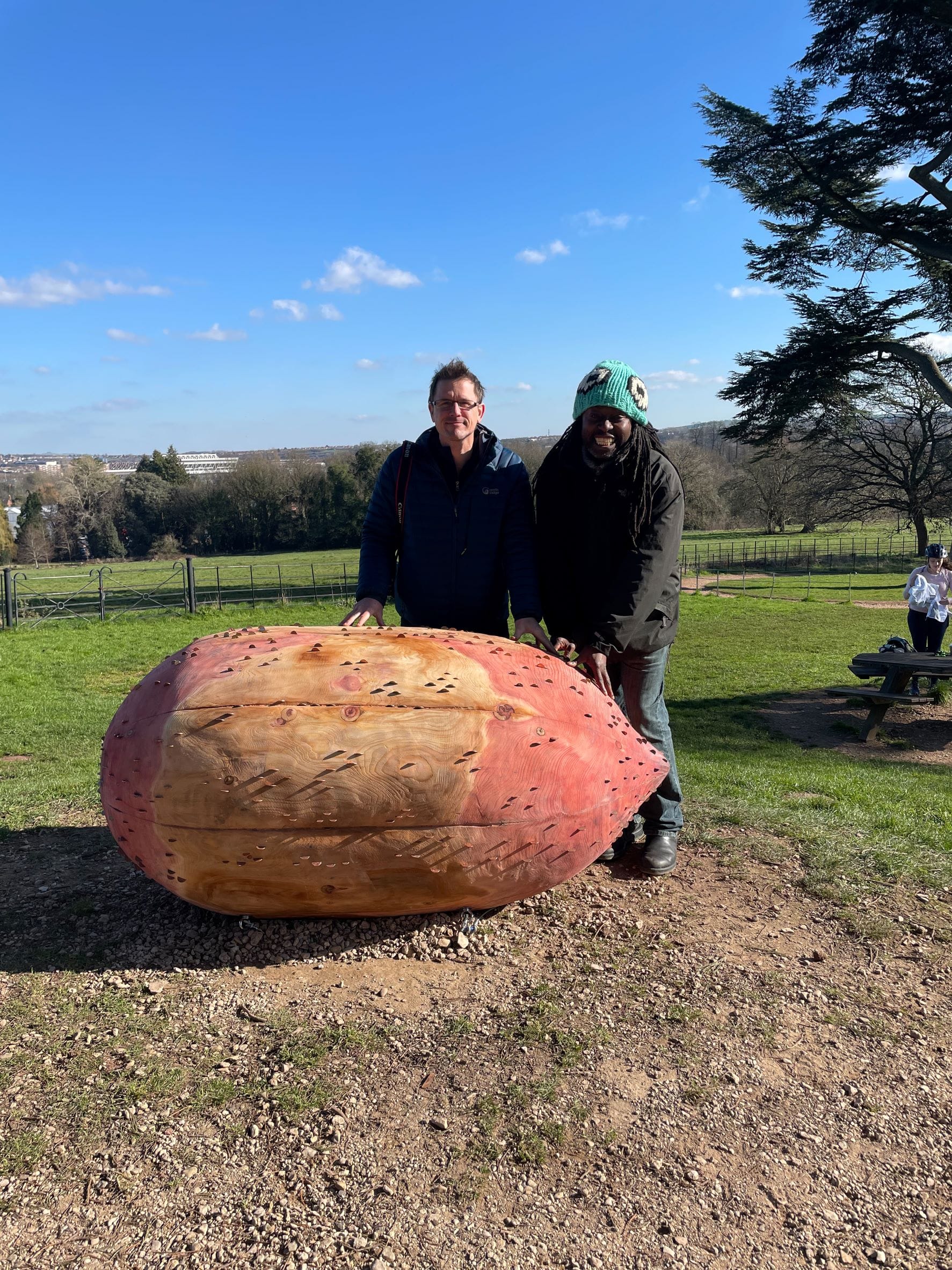 Two mnn are standing over a seed sculpture.  The man on the left is a white man, The man on the right is a black man wearing a green hat with eyes on it