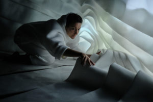 A woman reaches forwards surrounded by folds of white fabric