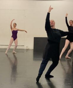 Three ballet dancers in a studio stand in plie with one arm raised