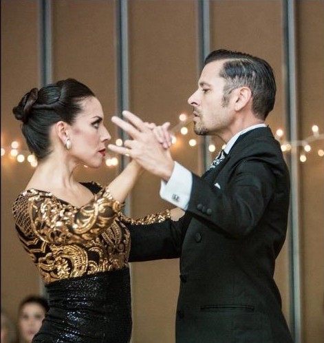 A smartly dressed couple of tango dancers stand in hold.
