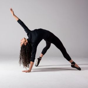 A dancer with long heair wearing black leggings, top and ballet shoes leans back on one arm in a white room