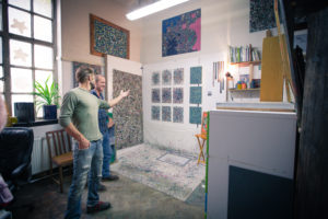 A man points to some artwork on the wall of his studio