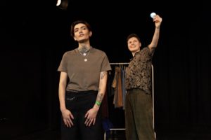 Two people stand on a stage in front of a clothes rail, one is taking a selfie