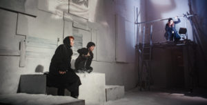 Two actors wearing warm winter clothes crouch on wooden blocks covered in fake snow.