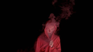 A person stands with a hood over their head which is filled with smoke