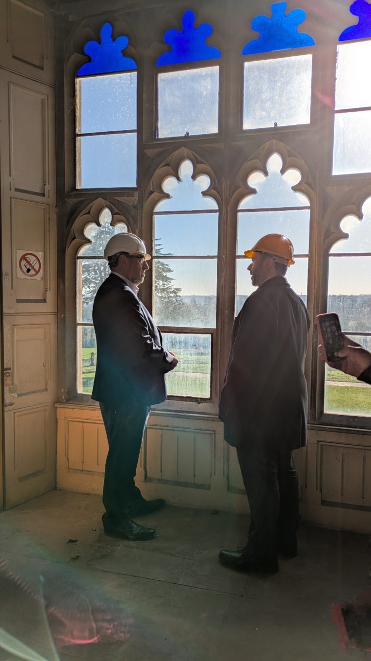 Two men wearing suits and hard hats stand in front of a sunlit ornate window in a mansion house.