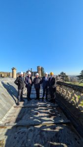 5 men are standing on a roof of a mansion house with an ornate stone parapet wearing suits and hard hats. Behind the men is a stair tower and views overlooking Bristol. The sky is sunny and clear..