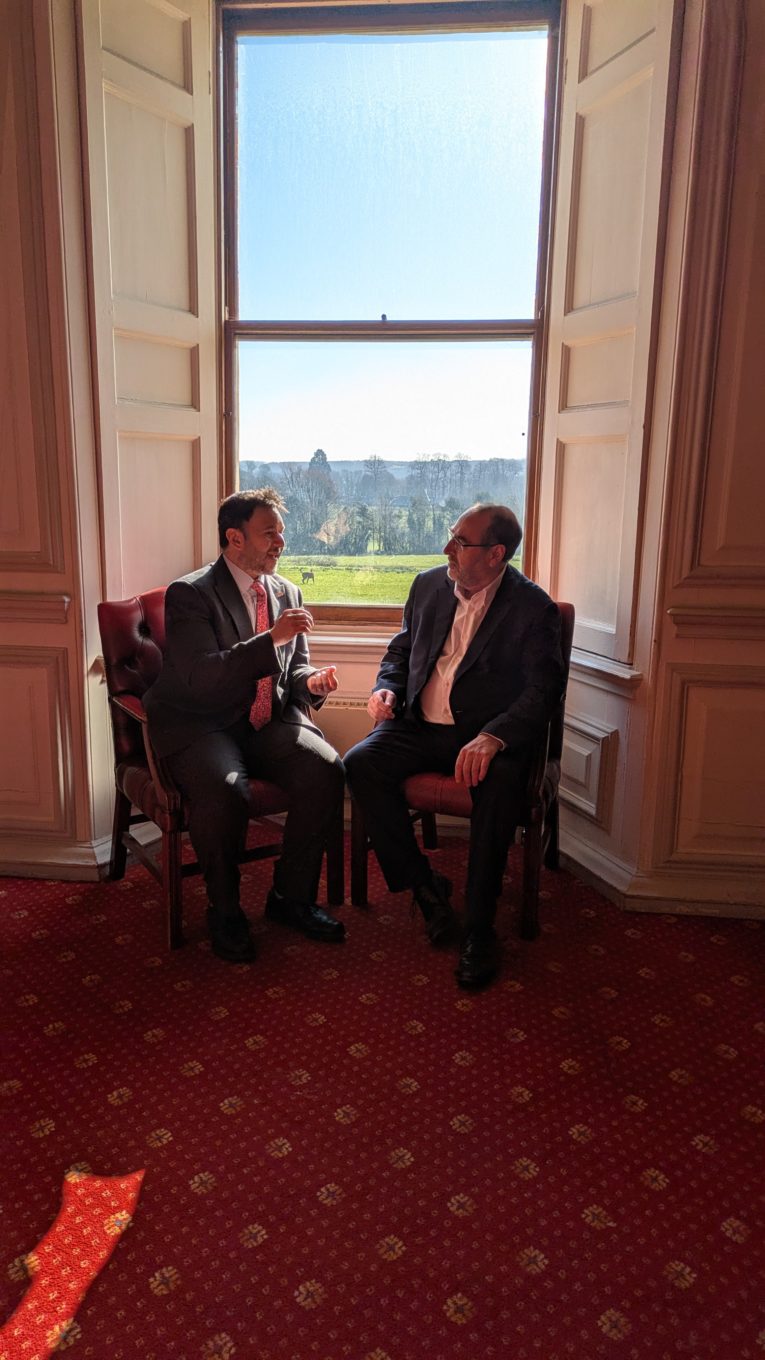 2 men sitting on chairs chatting. There is a window behind them from which you can see grass and trees. The chairs are leather and they are sitting facing each other.