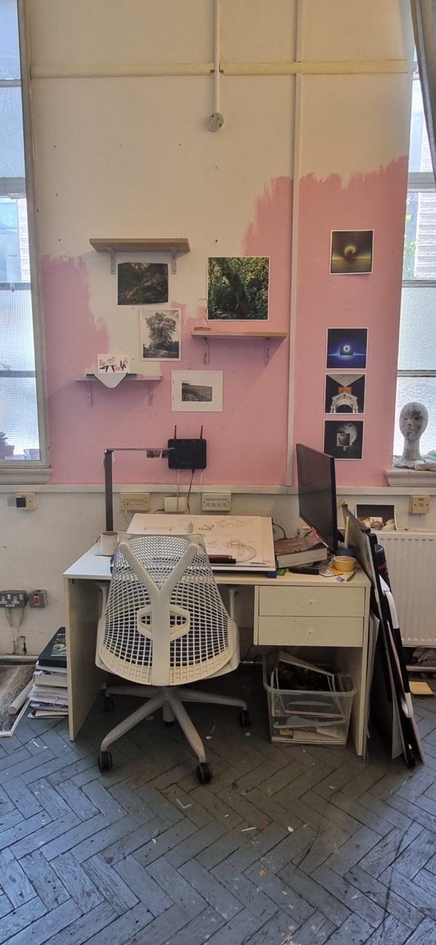 An artist studio desk space with a cluttered desk and, photos on the walls and a chair