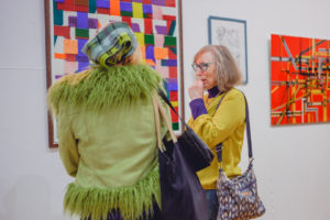 Two woman talk in front of some large artworks on a gallery wall.