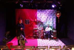 A theatre stage with two actors, one is sat on a sofa and one is standing at the front of the stage
