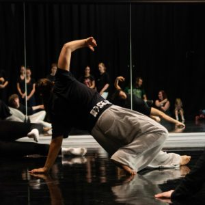 A dancer leaning on one hand with people watching them visable in a mirror