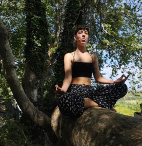 A woman sits on a low tree trunk with her legs crossed in a classic yoga pose