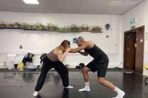 A man and a woman wrestle in a room with plants on a shelf