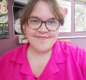 Rosa faces the camera and smiles. She is a white woman with brown hair and is wearing glasses and a bright pink shirt.