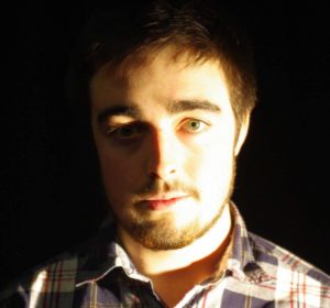 A man is standing lit againts a black background. He has dark hair and a beard and is wearing a check shirt.