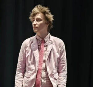 A man is standing against a black background. He has brown tousled hari and is wearing a pink shirt and jacket