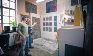 A man points to some artwork on the wall of his studio