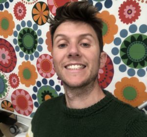 A white man grins a toothy grin in front of some flowery wallpaper