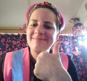 A white woman wearing a pink high viz tabard does a thumbs up to the camera