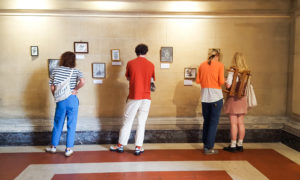 A group of four people stand in front of a wall with small framed artworks hanging on it