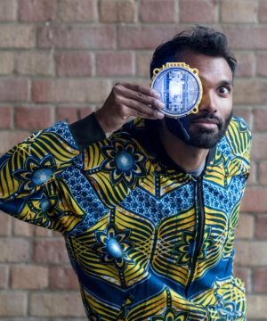 A man wearing a jazzy bomber jacket stars into the camera holding an intricate round light in front of one eye