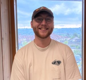 A man in a cap and t-shirt stands in front of a large window smiling at the camera