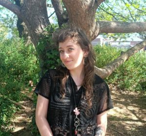 A white woman with long plaited hair stands by a tree in a forest